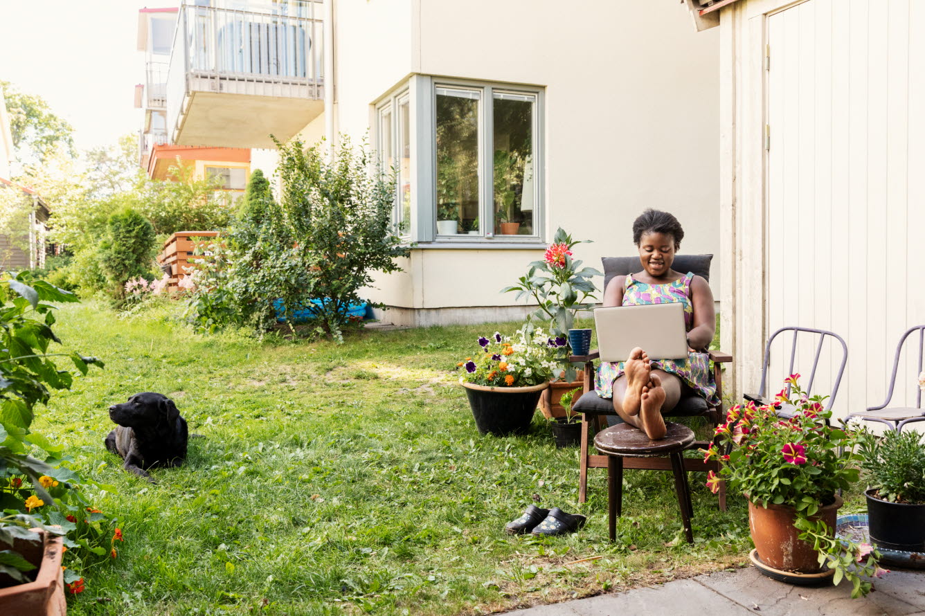 Kvinna med en laptop i knät i trädgårdsmiljö utanför flerfamiljshus.