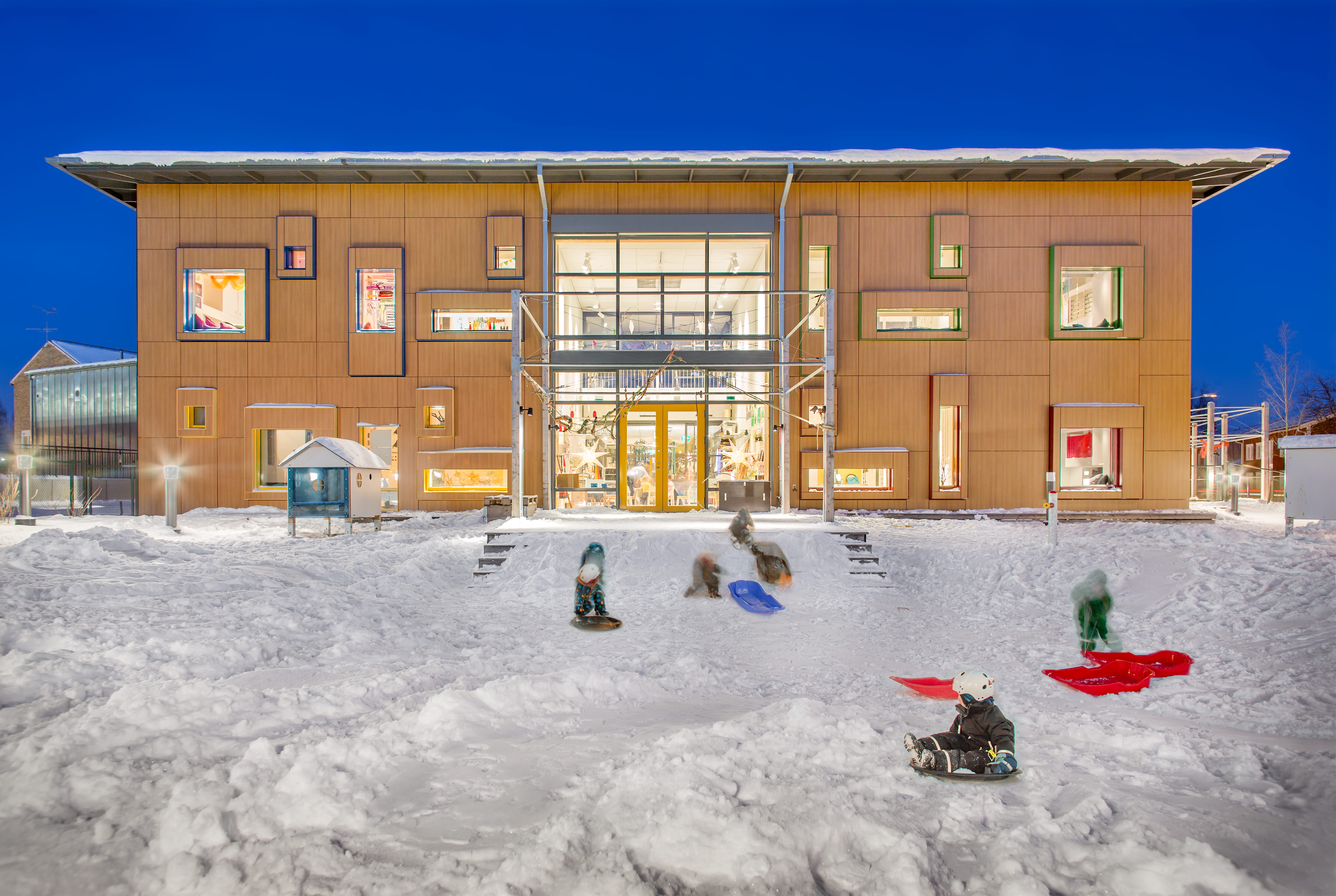 Snötäckt skolgård framför skolbyggnad som är ordentligt upplyst.