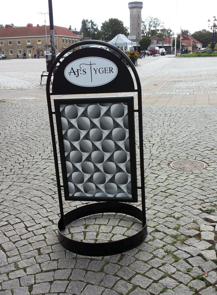 Foto på gatupratare i svart metall med ett grafiskt tyg och texten AJ:s Tyger stående på en gata.. 