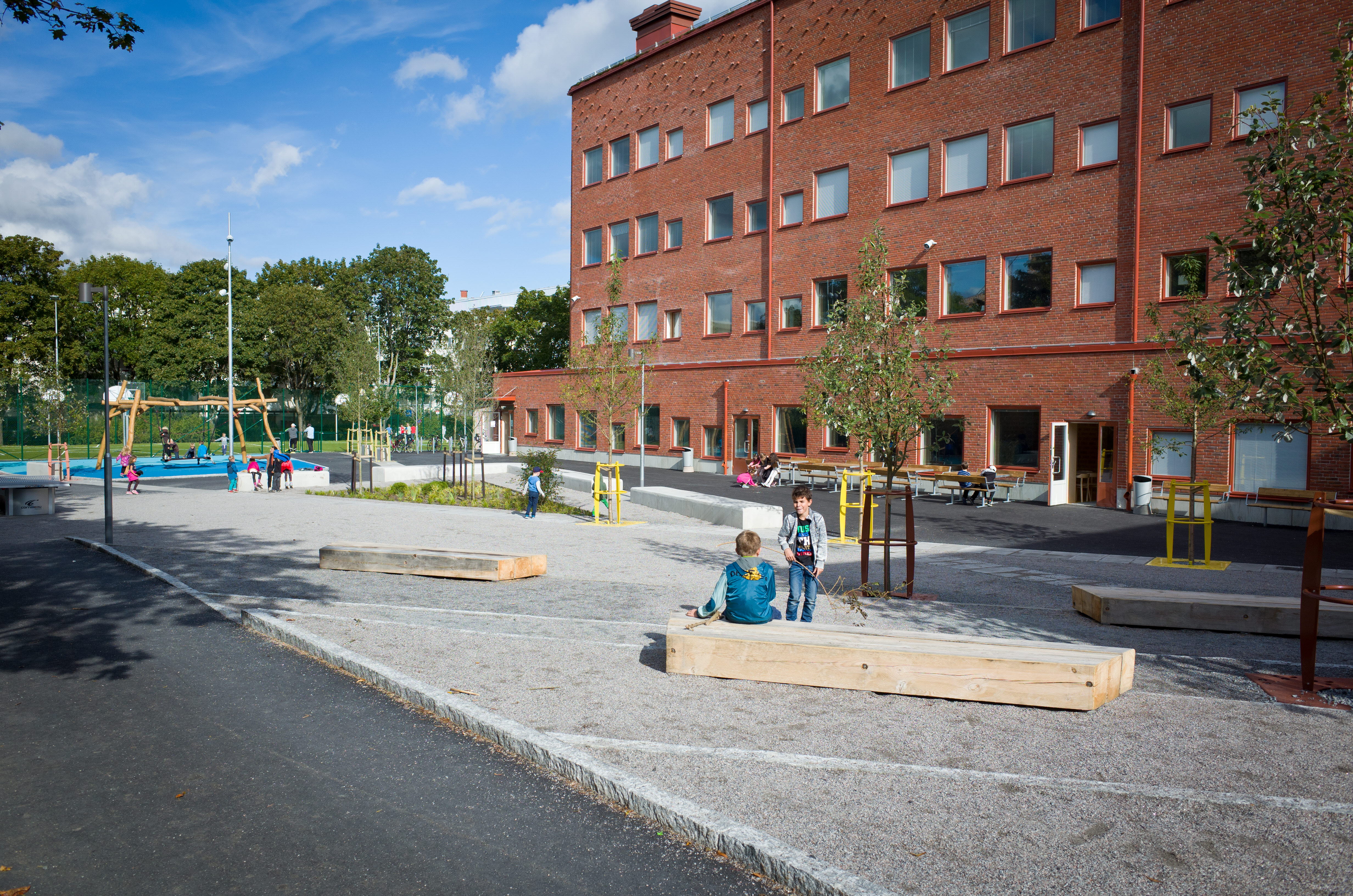 På marken ligger grus och det finns bänkar och små träd på skolgården. I bakgrunden står gungorna.