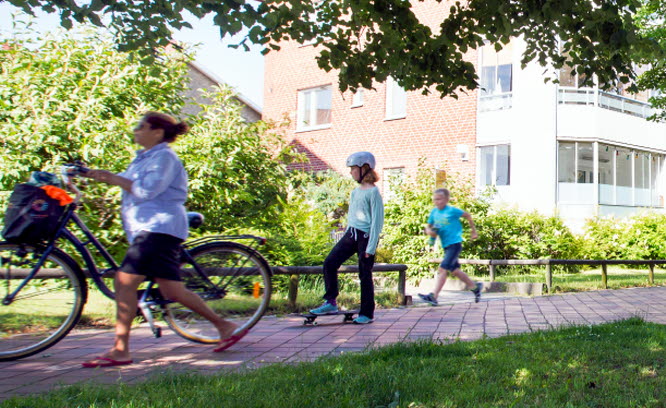 Cyklist, barn på skateboard och joggare på en trottoar framför ett flerbostadshus i en lummig miljö. Det är sommar.