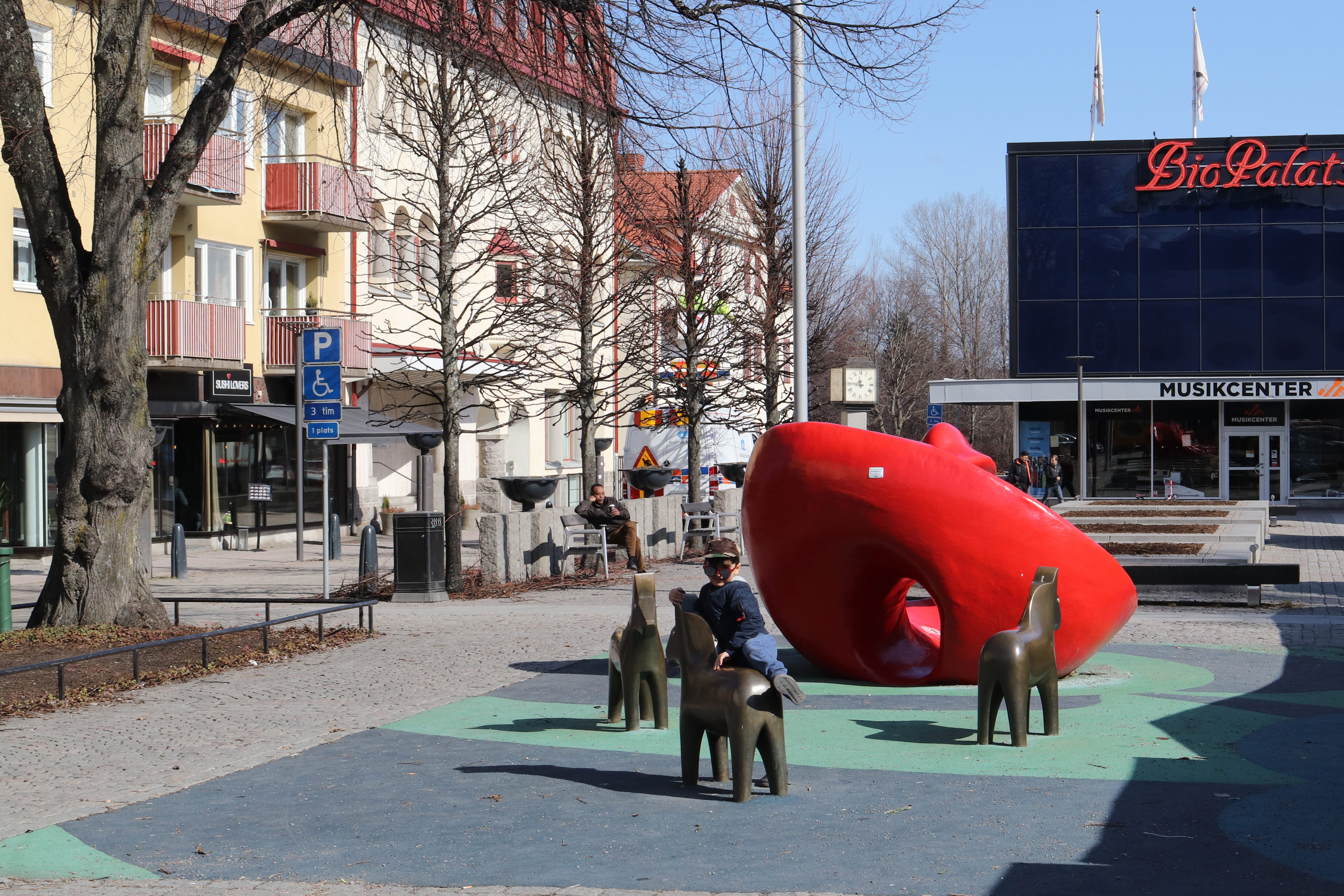 Lekplats på ett torg. Träd och byggnader runt torget. Lekredskapen består av dalahästar samt en stor röd rund skulptur. En pojke leker på lekplatsen.