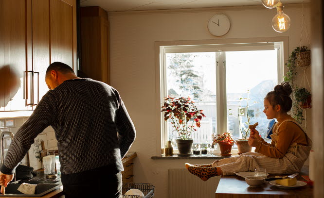 Pappa och flicka i ett kök. Pappan sätter in disk i diskmaskinen och flickan äter smörgås.