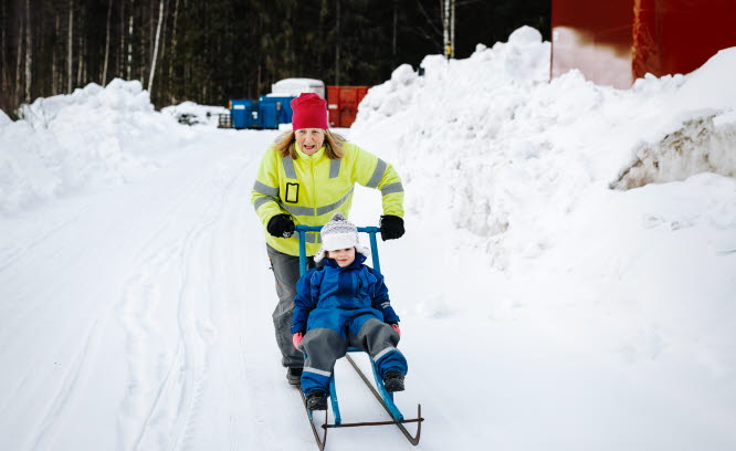 Kvinna i reflexjacka kör ett barn i overall på en spark på en snöig vinterväg.