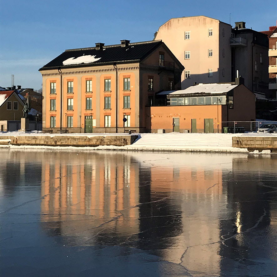 Några äldre putsade byggnader vid stranden speglar sig i en vattenyta med blank is