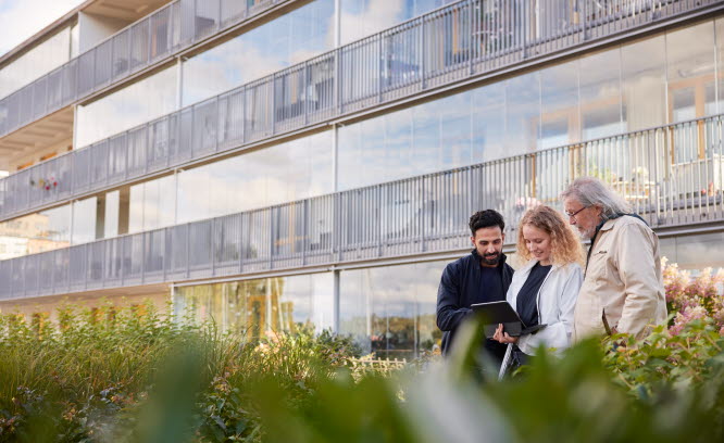 Tre personer står framför ett flerbostadshus och tittar på en skärm. Foto: Plattform/Scandinav Bildbyrå