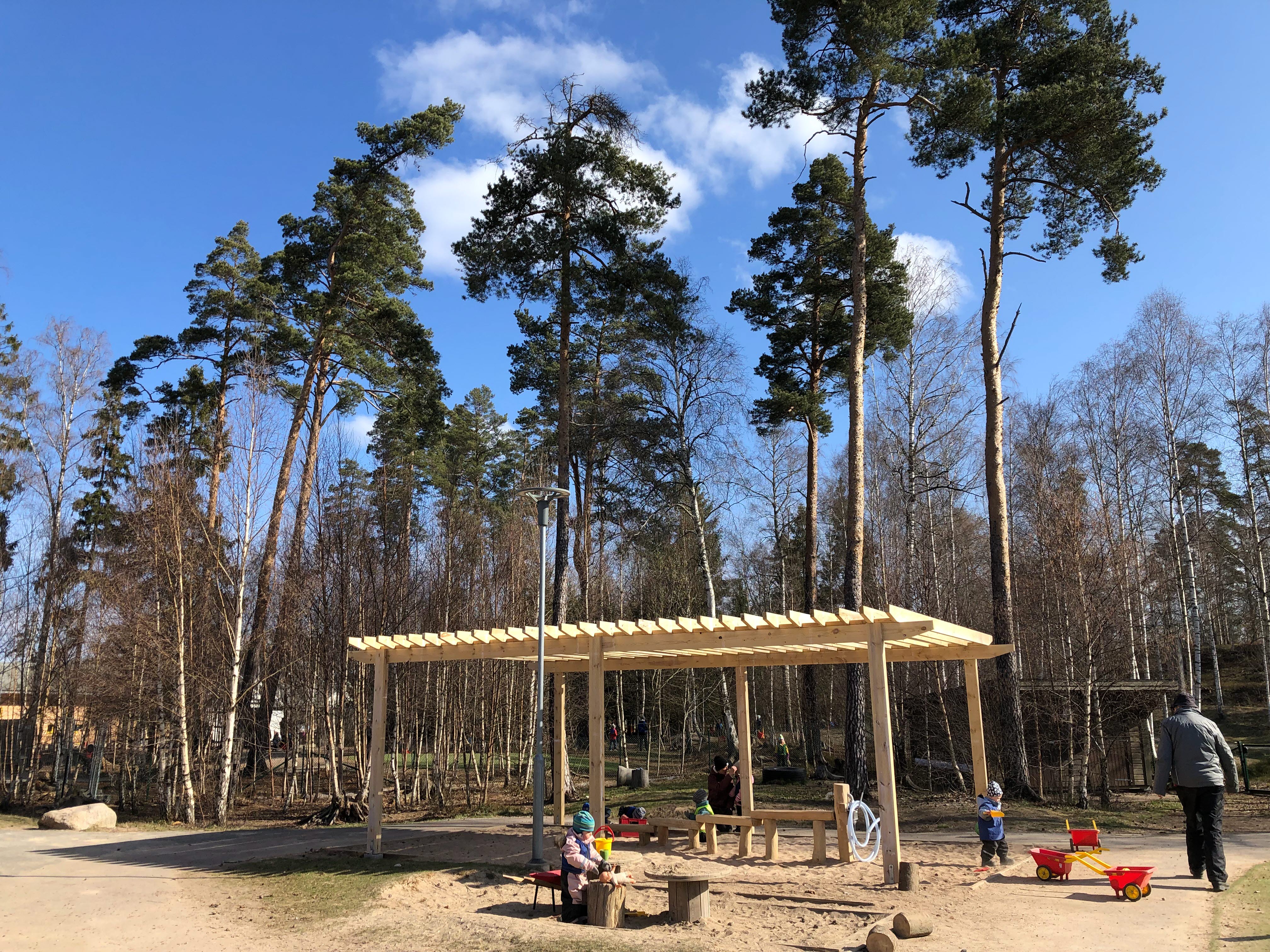 Foto med barn under ett solskydd i en sandlåda omgiven av höga tallar.