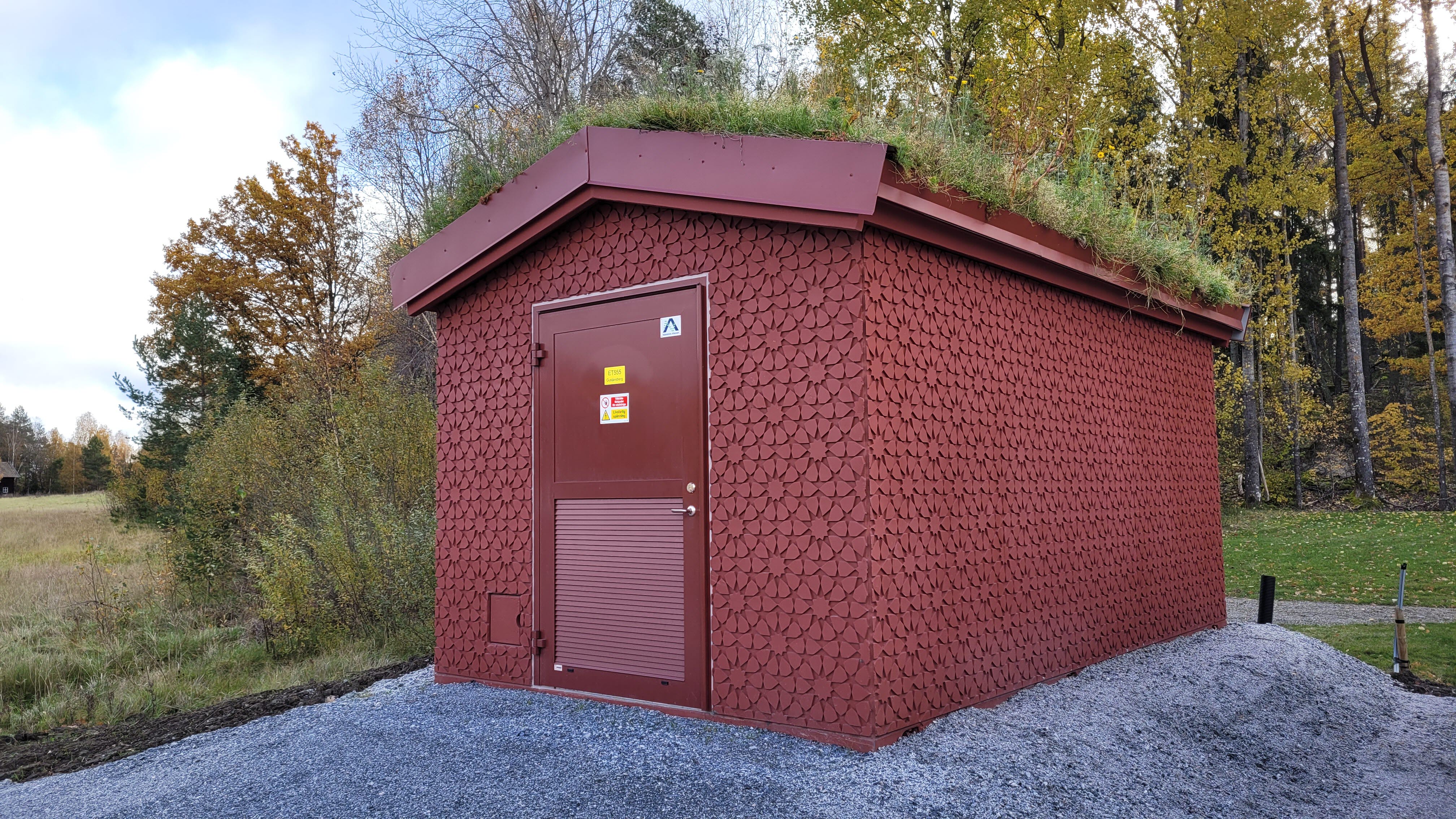 Foto av station med sluttande ängsbevuxet tak och blom-mönstrat fasadmaterial i faluröda nyanser.