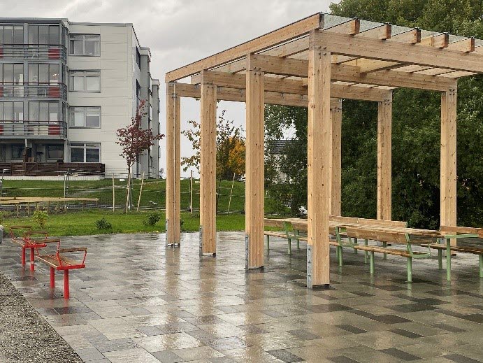 Pergola i trä med sittbänkar i trä under uppsatt på stenlagd plan.