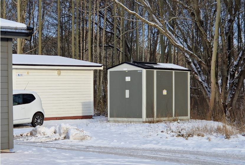 Foto på liten nätstation, placerad vid ett teknikhus i utkanten av ett bostadsområde.