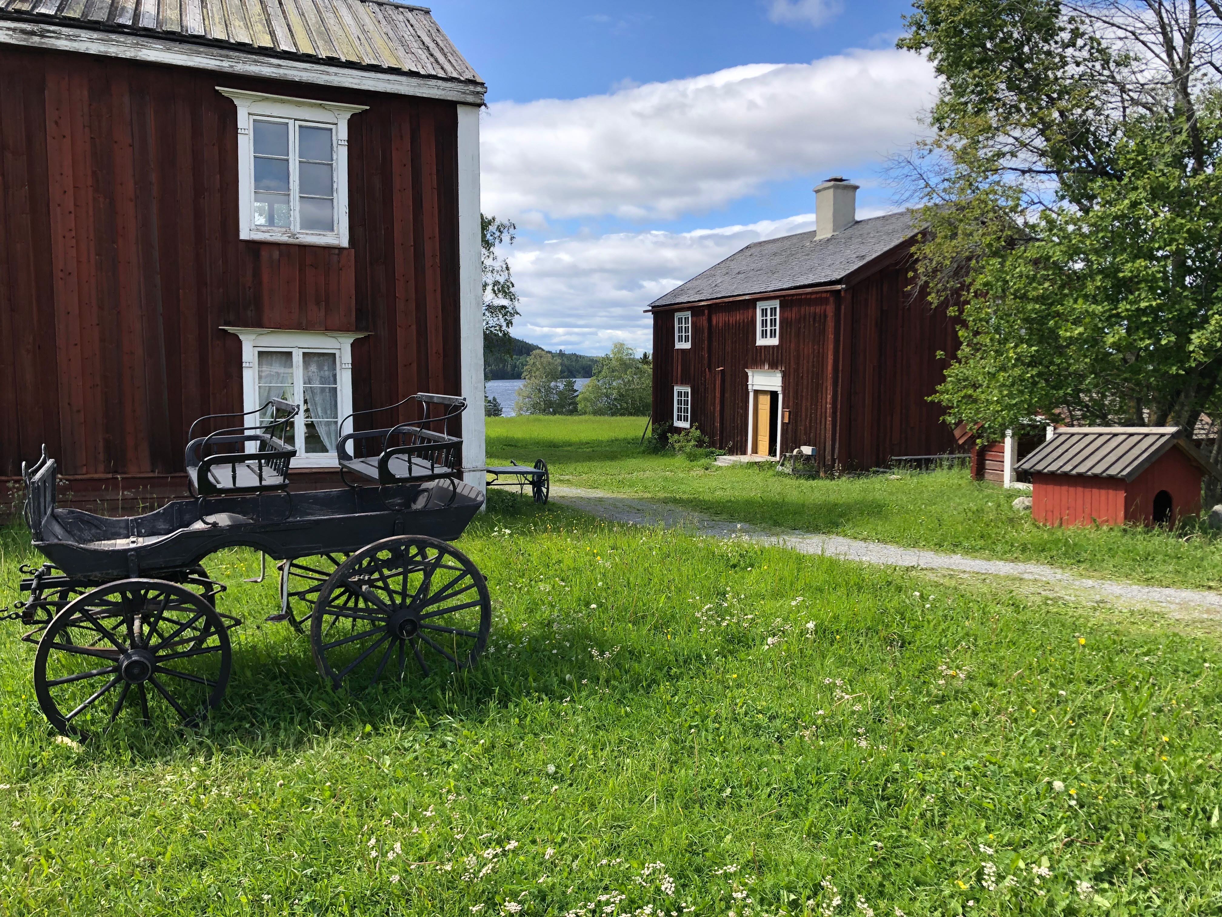 Röda kulturhistoriska byggnader, gräs, grusväg och en hästdroska.