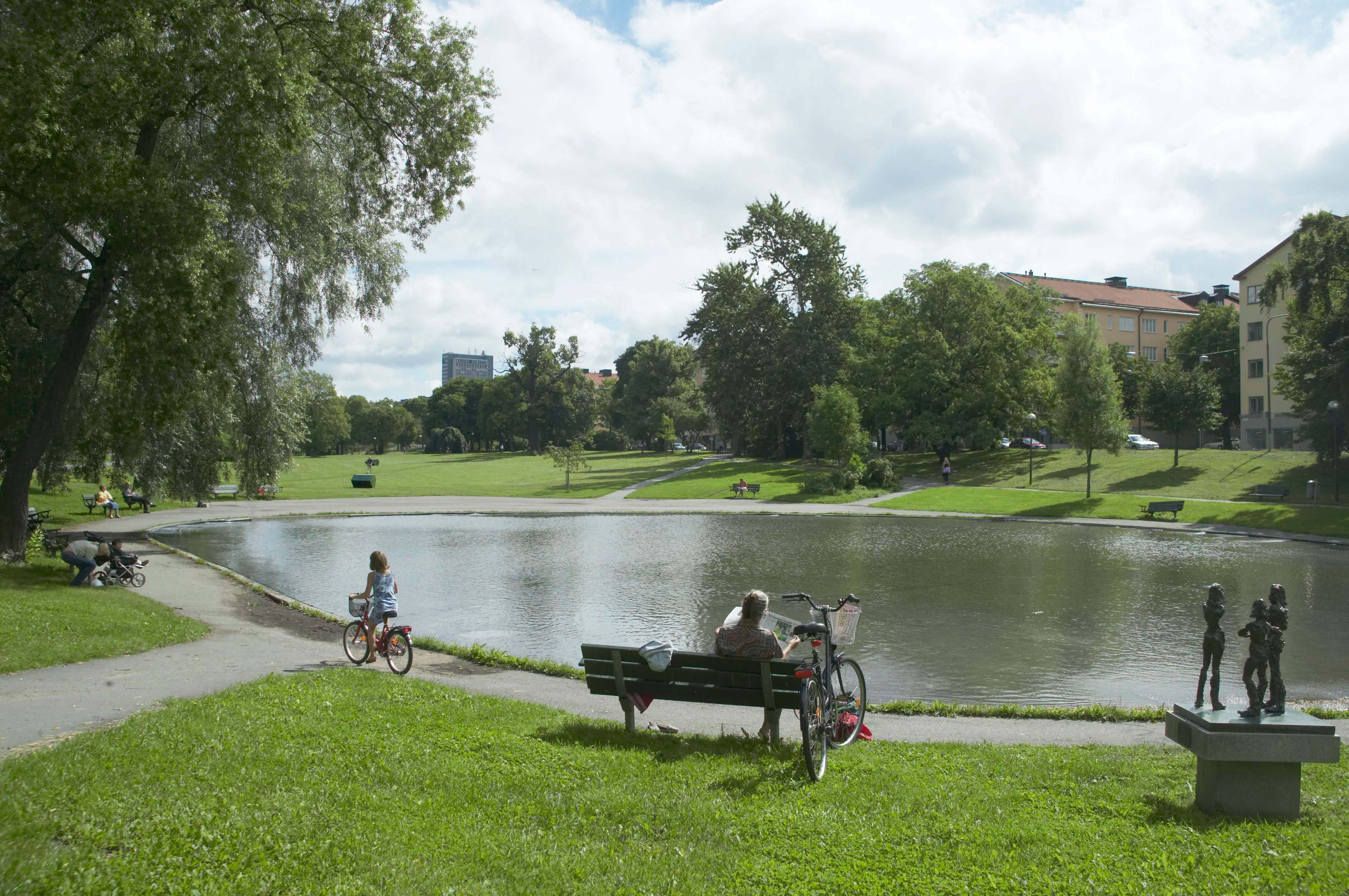 Park med en damm när människor sitter på bänkar och barn cyklar.