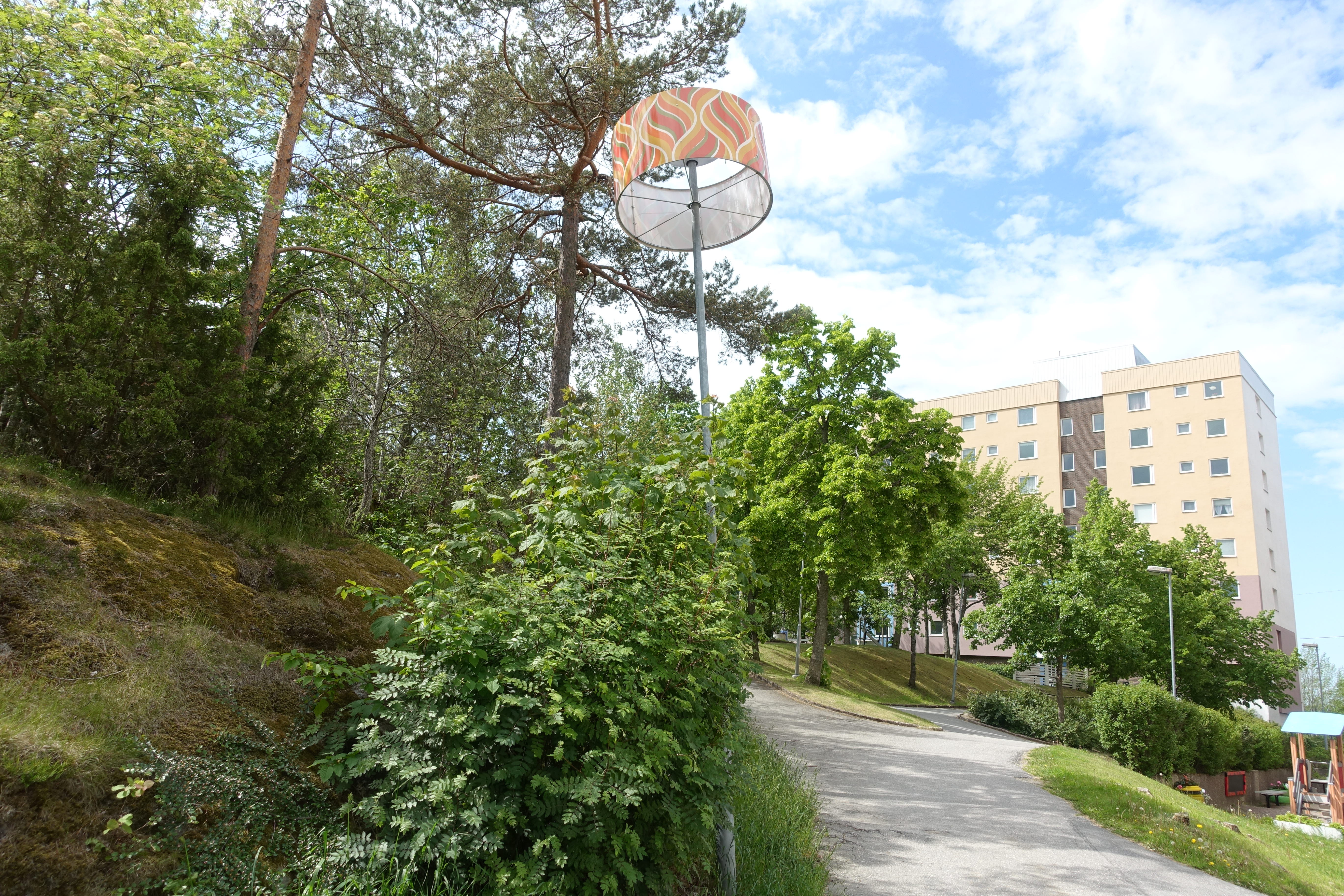 Lampa med lampskärm i park med flerbostadshus i bakgrunden.