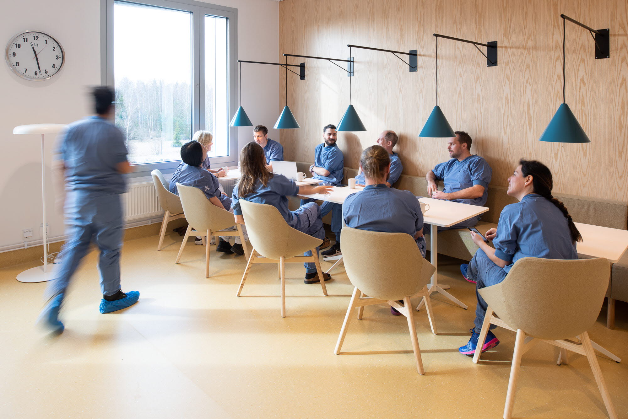 Blåklädd vårdperson på väg till ett konferensbord där flera andra blåklädda vårdpersoner sitter.