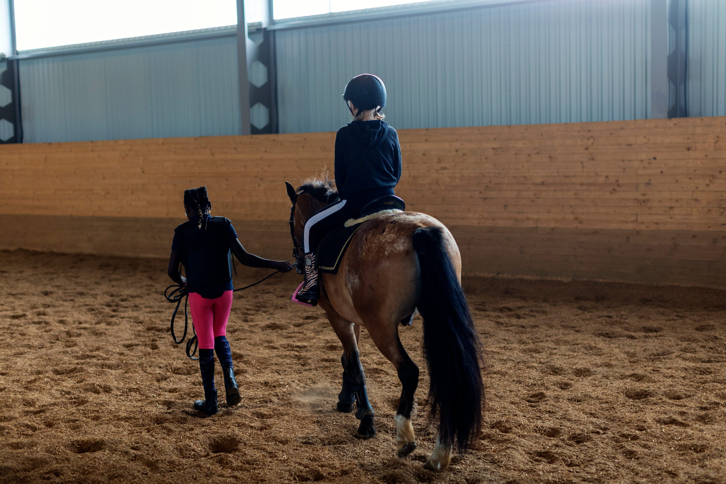 En person rider på en brun häst i ett ridhus. En person i röda byxor leder hästen