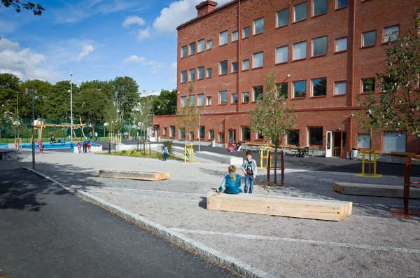 På marken ligger grus och det finns bänkar och små träd på skolgården. I bakgrunden står gungorna.