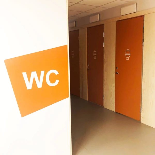 Toalettdörrar med en toalettsymbol. På väggen framför står WC på en orange färgplatta.