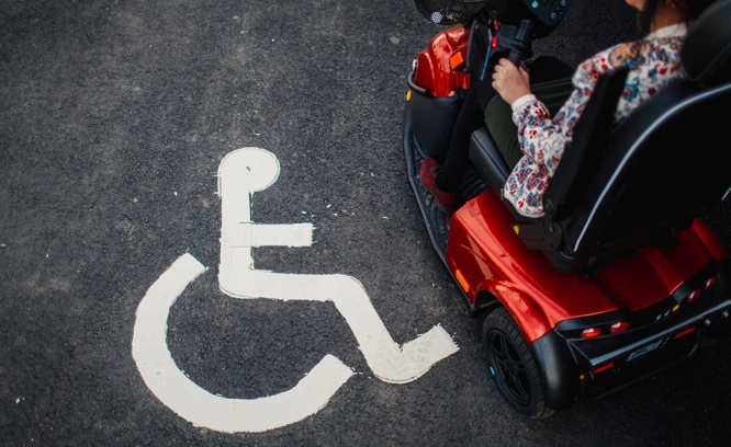 En person kör permobil bredvid en symbol för parkering för rörelsehindrade. Foto: Scandinav